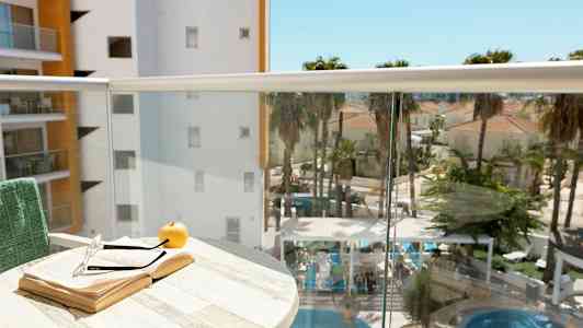 Parveke, jossa on avoin kirja, silmälasit ja omena pöydällä. Taustalla näkyy uima-allas ympäröivillä palmupuiden ja rakennusten kanssa. (Tekoälyn luoma teksti)