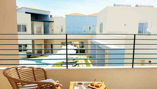 Näkymä parvekkeelta sisäpihalle, jossa on pieni uima-allas, ympäröitynä vaaleilla rakennuksilla. Parvekkeella on tuoli ja pöytä, jonka päällä on aurinkolasit ja kahvikuppi. (Tekoälyn luoma teksti)