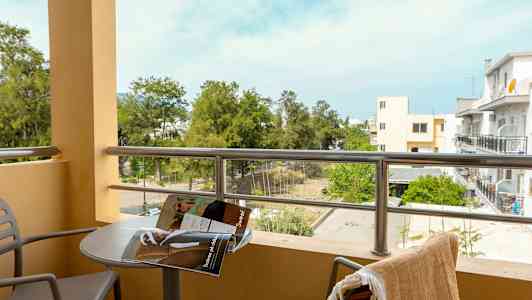 Parveke, josta näkymä vehreyteen ja rakennuksiin, pöytä lehden kanssa ja kaksi tuolia. (Tekoälyn luoma teksti)