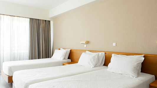 Kaksi sänkyä, joissa valkoiset peitot ja tyynyt, valoisassa huoneessa, jossa verhot. (Tekoälyn luoma teksti)