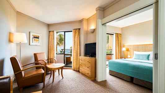 Hotellihuone, jossa istuinryhmä, televisio ja sänky sinisellä päiväpeitteellä. (Tekoälyn luoma teksti)