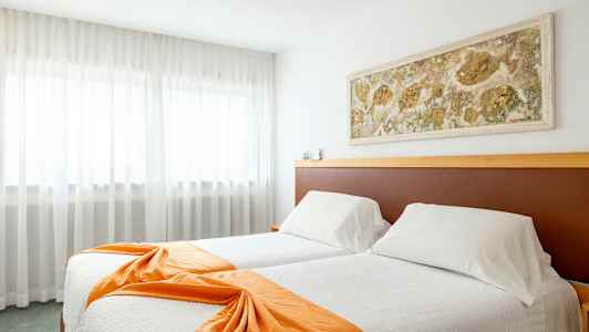 Hotellihuone, jossa on kaksi sänkyä valkoisin vuodevaattein ja oranssein koristehuivein, seinäkoriste sänkyjen yläpuolella sekä verhot suuren ikkunan edessä. (Tekoälyn luoma teksti)