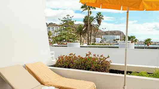 Aurinkotuoli, jossa pyyhe parvekkeella näkymällä palmuihin ja rakennuksiin. (Tekoälyn luoma teksti)