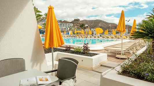 Terassi, jossa kalusteet, näkymät uima-allasalueelle keltaisine aurinkovarjoineen. (Tekoälyn luoma teksti)