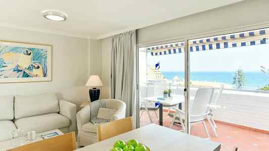 Olohuoneen sisätila, jossa sohva, nojatuoli ja pöytä, sekä näkymä merelle lasiovien kautta terassille. (Tekoälyn luoma teksti)