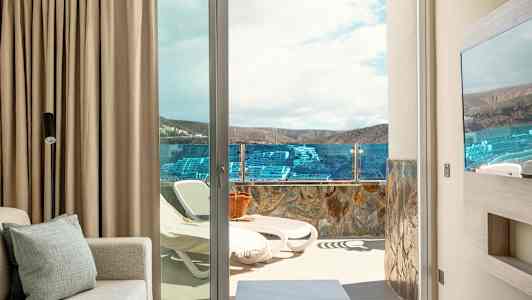 Huoneesta näkymä terassille, jossa aurinkotuoleja ja lasikaide, taustalla maisema ja taivas. (Tekoälyn luoma teksti)