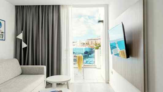 Olohuone, jossa sohva, pöytä, televisio ja avoin parvekkeen ovi, josta näkyvät rakennukset ja meri. (Tekoälyn luoma teksti)