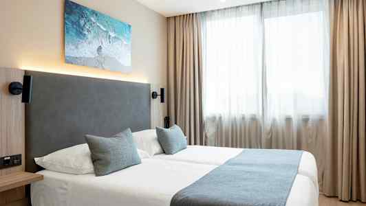 Hotellihuone, jossa on kaksi vuodetta, siniset vuodevaatteet ja kaksi tyynyä ikkunoiden edessä, joissa on vaaleat verhot. (Tekoälyn luoma teksti)