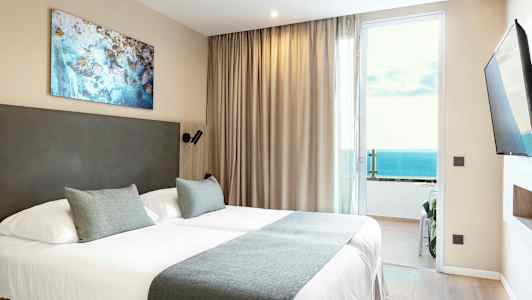 Hotellihuone, jossa on parivuode, beigejä verhoja ja merinäköala lasioven kautta. (Tekoälyn luoma teksti)