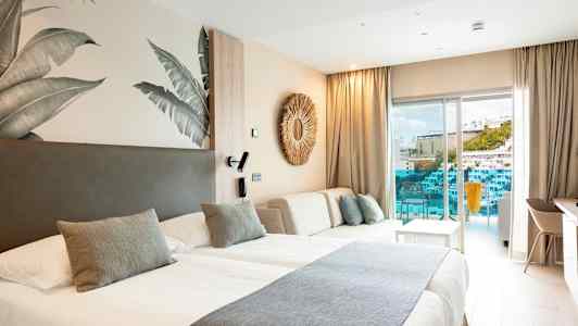 Moderni hotellihuone, jossa on kaksi sänkyä, vaalea sisustus ja parveke näköalalla uima-altaalle ja rakennuksiin. (Tekoälyn luoma teksti)