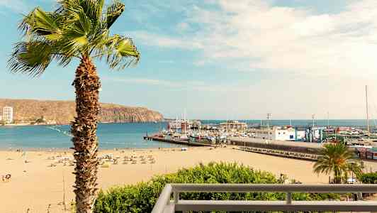 Ranta aurinkotuoleineen ja aurinkovarjoineen, satama veneineen ja palmu etualalla. (Tekoälyn luoma teksti)