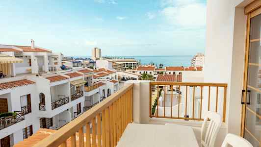 Näkymä parvekkeelta, jossa on pöytä ja tuolit, valkoisten rakennusten yli punaisine kattoineen ja taustalla meri. (Tekoälyn luoma teksti)