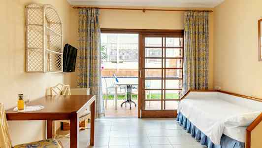 Hotellihuone, jossa on yhden hengen sänky, pöytä ja tuolit, sekä näkymä huonekalulla varustetulle terassille. (Tekoälyn luoma teksti)