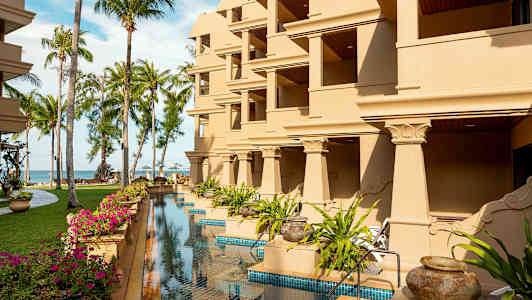 Rakennus, jossa on useita parvekkeita, ympäröity nurmikoilla, palmuilla ja uima-altaalla. (Tekoälyn luoma teksti)