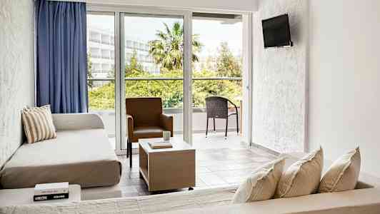 Valoisa hotellihuone, jossa on sohvia, pöytä, tuoli ja parveke ulkopuolella. (Tekoälyn luoma teksti)