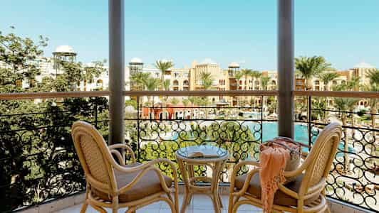 Näköala parvekkeelta, jossa on kaksi rottinkituolia ja pöytä, uima-altaalle ja hotellille, joita ympäröivät palmut. (Tekoälyn luoma teksti)