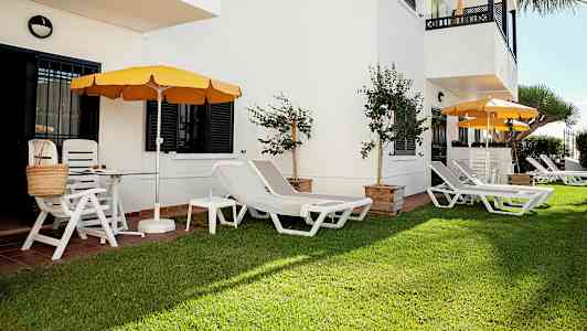 Vihreä alue, jossa aurinkotuoleja ja aurinkovarjoja valkoisen rakennuksen vieressä. (Tekoälyn luoma teksti)