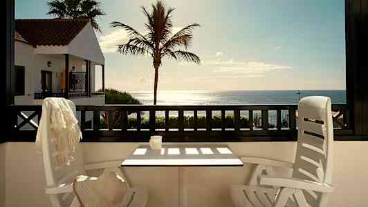 Näkymä terassilta, jossa on pöytä ja tuolit, kohti merta. Taustalla palmu ja punakattoinen rakennus. (Tekoälyn luoma teksti)