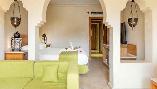 Hotellihuone, jossa sänky koristeltu muotoillulla pyyhkeellä ja kukilla, vihreä sohva etualalla ja koristeelliset valaisimet. (Tekoälyn luoma teksti)