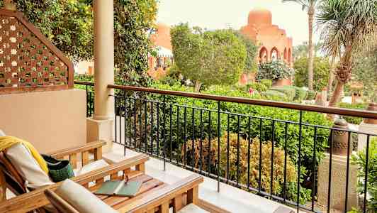 Terassi, jossa on puiset tuolit ja näkymä vehreään puutarhaan ja rakennuksiin, joissa on kaari-ikkunoita. (Tekoälyn luoma teksti)