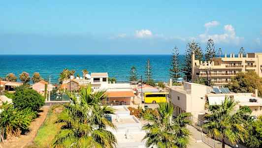 Näkymä asuinalueelle, jossa palmuja ja rakennuksia, ja taustalla näkyy meri kirkkaan sinisen taivaan alla. (Tekoälyn luoma teksti)