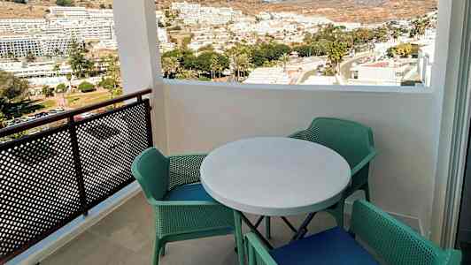 Näkymä parvekkeelta, jossa on pyöreä pöytä ja neljä vihreää tuolia, näkymä kaupunkiin ja vuorille taustalla. (Tekoälyn luoma teksti)