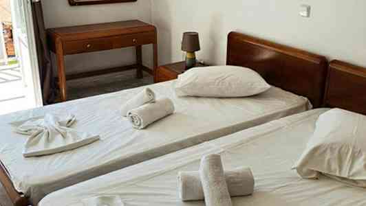 Kaksi yhden hengen sänkyä valkoisilla lakanoilla ja pyyhkeet asetettuna päälle, sekä pöytä ja peili huoneessa. (Tekoälyn luoma teksti)