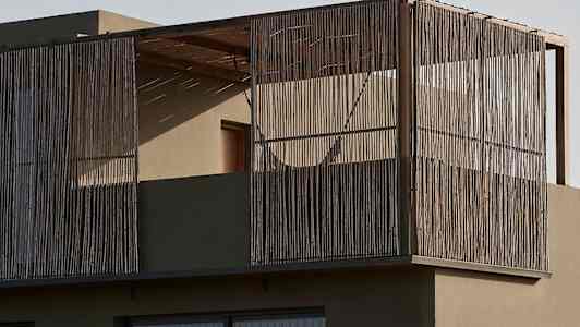 Nykyaikainen rakennus, jossa parveke on ympäröity puutangoilla ja lasi-ikkunoilla. Edessä näkyy kasvillisuutta aavikkomaisemassa. (Tekoälyn luoma teksti)