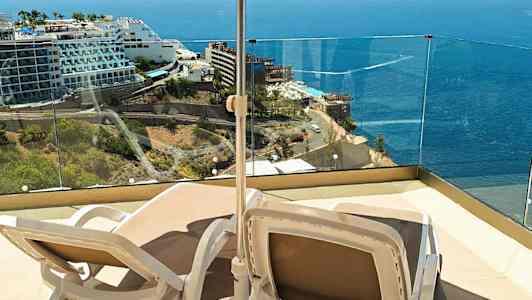 Kaksi aurinkotuolia varjon alla terassilla, josta on näkymä merelle ja läheisiin hotelleihin. (Tekoälyn luoma teksti)