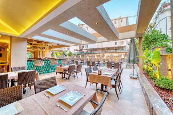 Ulkoterassi, jossa pöydät aseteltu astioilla ja ruokailuvälineillä sekä taustalla vihreitä kasveja ja uima-allas. (Tekoälyn luoma teksti)