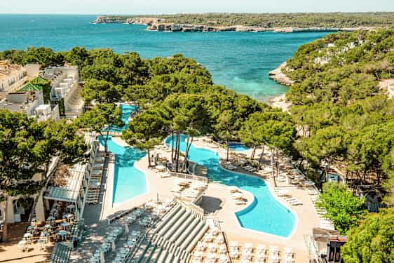 Näkymä lomakeskuksesta, jossa on uima-altaita aurinkotuolien ja aurinkovarjojen reunustamina, sijaitsee turkoosin meren äärellä vehreän kasvillisuuden keskellä. (Tekoälyn luoma teksti)