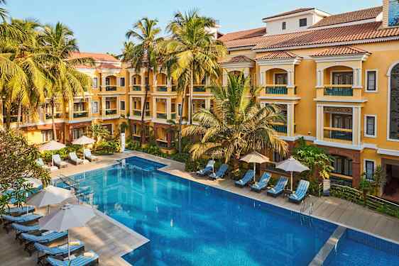 Kuvassa hotellirakennus, jossa on keltainen julkisivu ja punainen katto, ympäröitynä palmupuiden kanssa. Rakennuksen edessä on suuri uima-allas ja aurinkotuoleja sateenvarjoilla. (Tekoälyn luoma teksti)