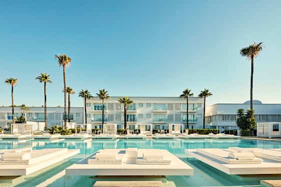 Moderni rakennus, jossa parvekkeet, ympäröity palmilla ja uima-altaalla, jossa on aurinkotuoleja. (Tekoälyn luoma teksti)