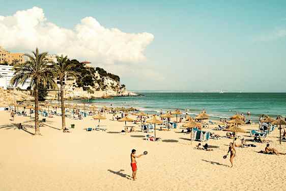 Hiekkaranta, jossa on aurinkovarjoja, aurinkotuoleja ja ihmisiä nauttimassa auringosta ja merestä. Kauempana näkyy kallio ja rakennuksia. (Tekoälyn luoma teksti)