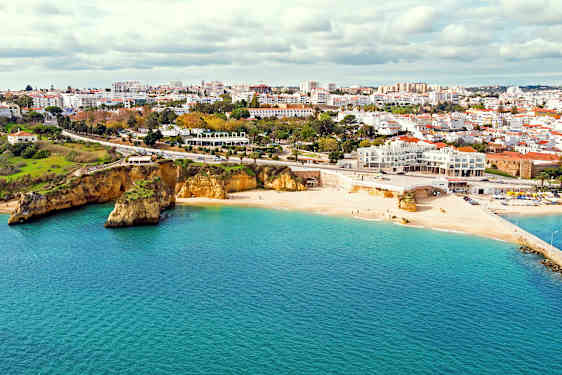 Vaalea hiekkaranta meren äärellä, kaupungin ollessa taustalla ja kallioiden ranta-alueella. (Tekoälyn luoma teksti)