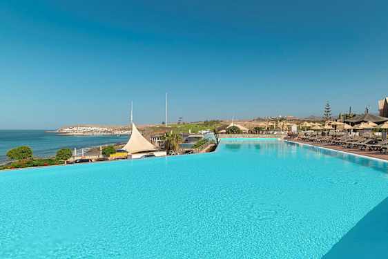 Suuri ulkouima-allas, jonka vieressä on aurinkotuoleja ja varjokatos. Näköala merelle ja rannikkokaupunkiin. (Tekoälyn luoma teksti)