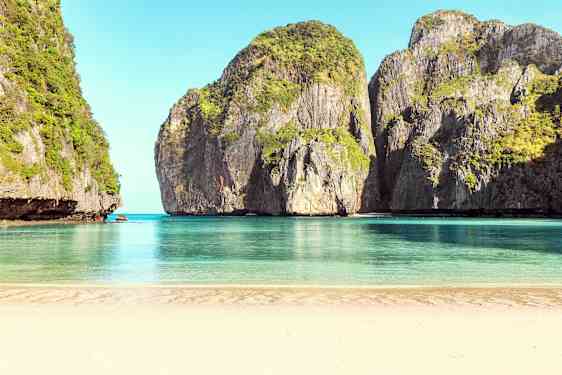 Hiekkaranta, joka avautuu rauhalliseen, siniseen veteen, ympäröitynä korkeilla kasvillisuuden peittämillä kallioilla. (Tekoälyn luoma teksti)