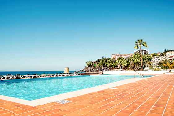 Uima-allas ympäröitynä aurinkotuoleilla, näkymä merelle ja rakennuksia palmujen keskellä. (Tekoälyn luoma teksti)