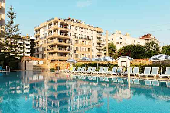 Kuva uima-altaasta, jonka sivulla aurinkotuoleja ja aurinkovarjoja, taustalla rakennus. (Tekoälyn luoma teksti)