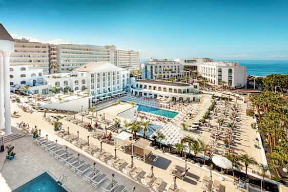 Näkymä hotellialueelle, jossa on uima-altaita, aurinkotuoleja ja vaalea arkkitehtuuri meren lähellä. (Tekoälyn luoma teksti)