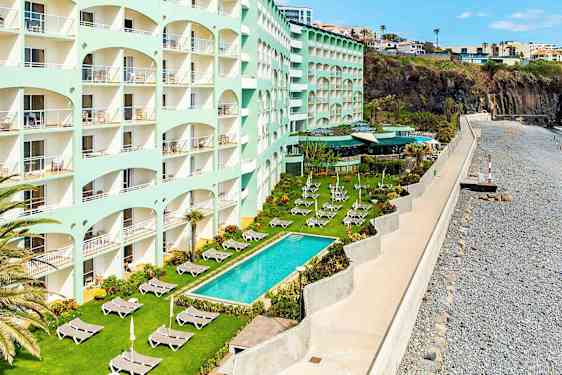 Hotelli, jossa on useita kerroksia meren äärellä, nurmikko aurinkotuoleilla ja uima-allas ulkona. (Tekoälyn luoma teksti)