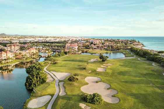 Golfkenttä, jossa on hiekkabunkkereita ja vihreitä nurmialueita lähellä vesiväylää ja rantaa. Taustalla rakennuksia. (Tekoälyn luoma teksti)