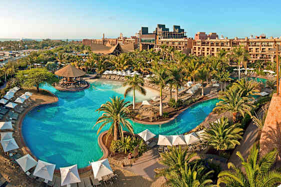 Suuri ulkoallas, jonka ympärillä on varjoja, aurinkotuoleja ja vehreää kasvustoa lomahotellissa. (Tekoälyn luoma teksti)