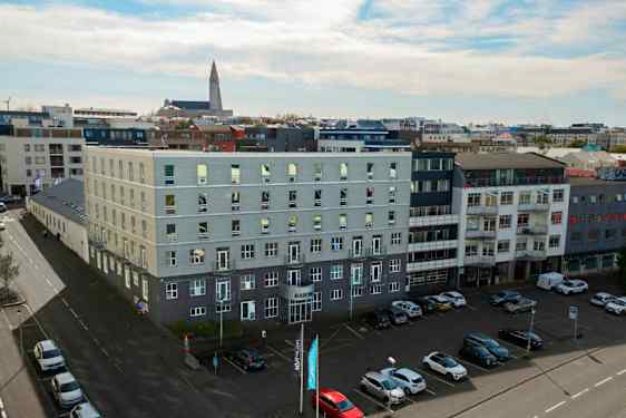 Useat modernit rakennukset ympäröivät avointa pysäköintialueita, jossa on autoja pysäköitynä, ja kirkko näkyy taustalla. (Tekoälyn luoma teksti)
