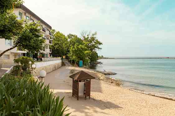 Rakennus, jossa on useita parvekkeita, hiekkaranta ja meri puiden kanssa sekä etualalla lehtimaja. (Tekoälyn luoma teksti)