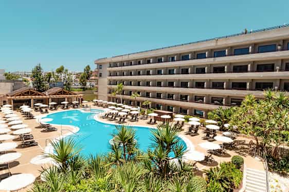 Hotellirakennus parvekkeineen ja uima-allas, jota ympäröivät aurinkotuolit ja aurinkovarjot. (Tekoälyn luoma teksti)