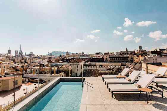 Ulkoallas ja aurinkotuoleja kattoterassilla, josta on näkymä yli kaupungin rakennusten, kirkkaalla taivaalla. (Tekoälyn luoma teksti)