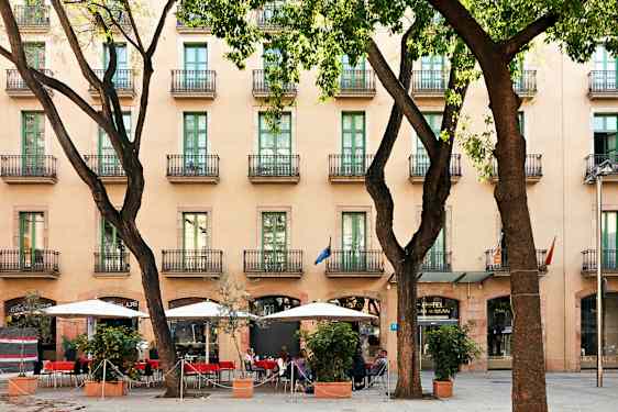 Hotelli, jossa on useita kerroksia ja parvekkeita, jalkakäytävä, jolla on aurinkovarjoja ja istumapaikkoja puiden joukossa. (Tekoälyn luoma teksti)