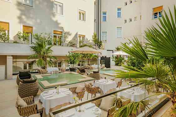 Kodikas sisäpiha, jossa on tyylikkään ympäristön pöytiä ja tuoleja. Siellä on myös vihreitä kasveja ja pieni allas. (Tekoälyn luoma teksti)