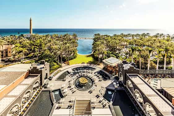 Näkymä hotellin puutarhaan, jossa palmuja, suuri uima-allas ja meri taustalla. (Tekoälyn luoma teksti)